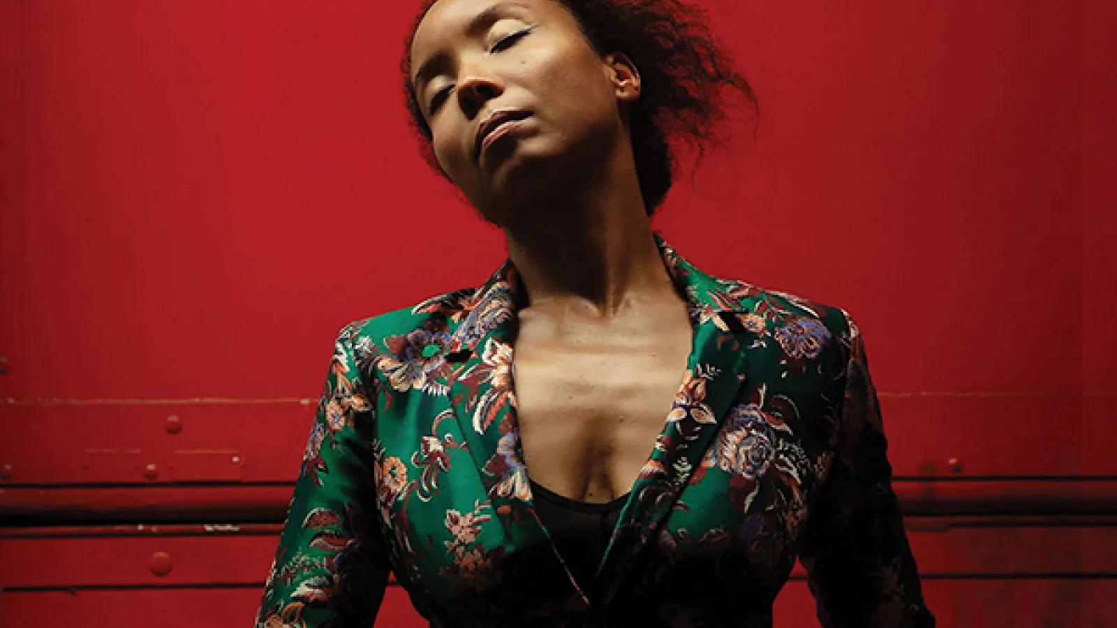 Person with eyes closed wearing green patterned dress in front of red background