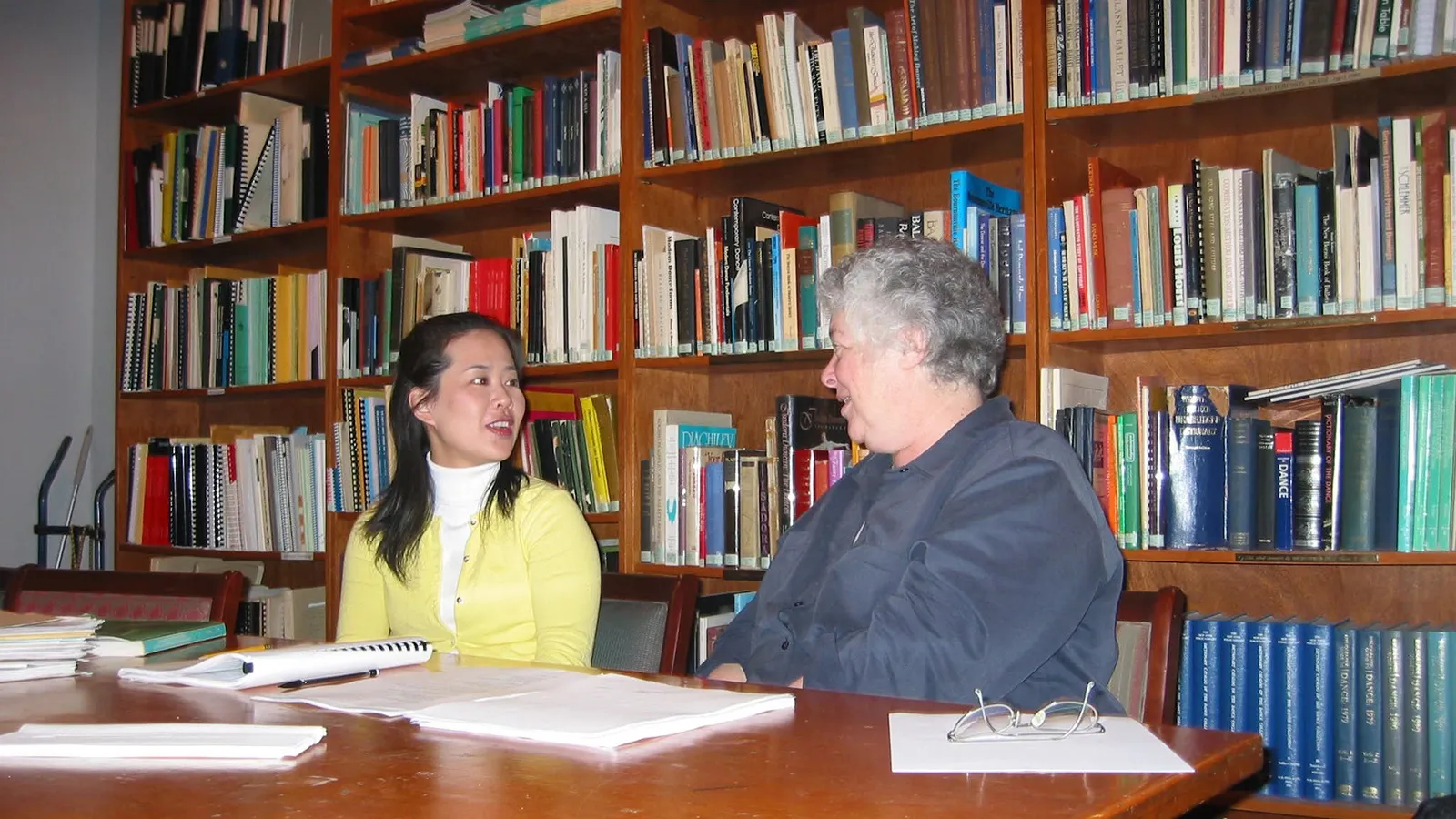 two people talking at a table in front of a bookshelf