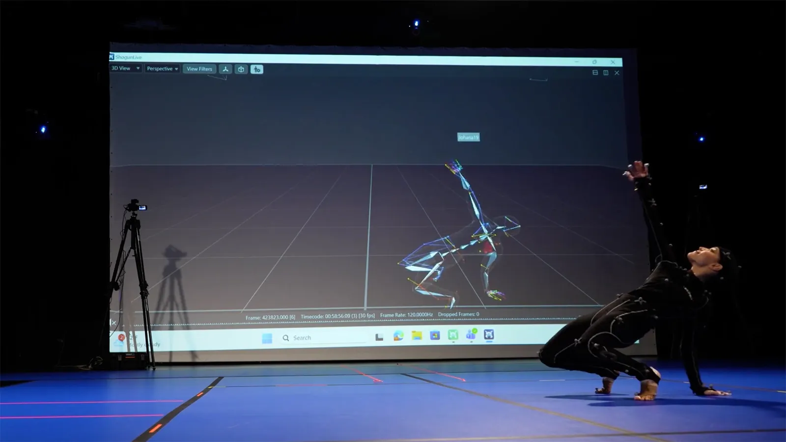 A dancer wearing a motion capture suit moving in front of a screen with her avatar projected on it