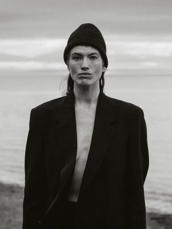 black and white photo of a woman in a black blazer and hat in front of the ocean 